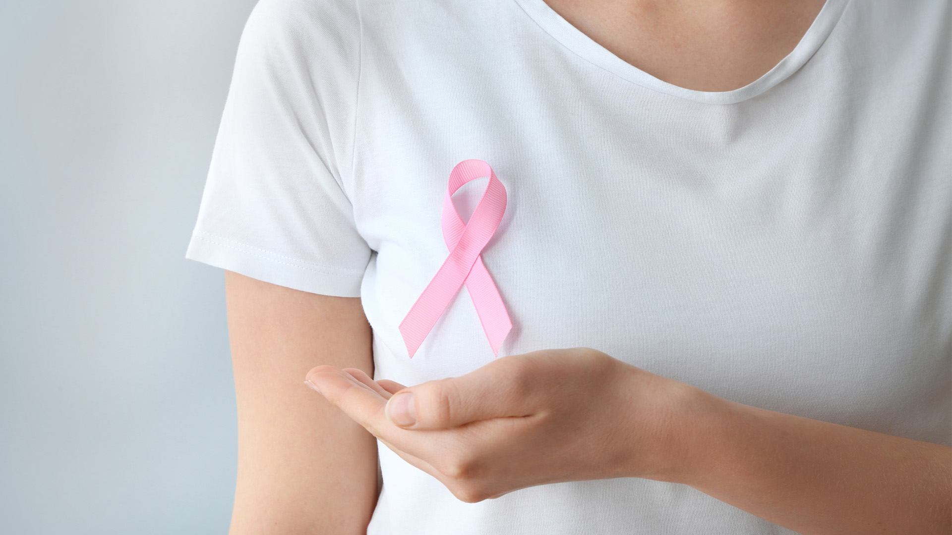 Frau hält Hand auf Brusthöhe und darüber symbolisch eine rosafarbene Krebsschleife